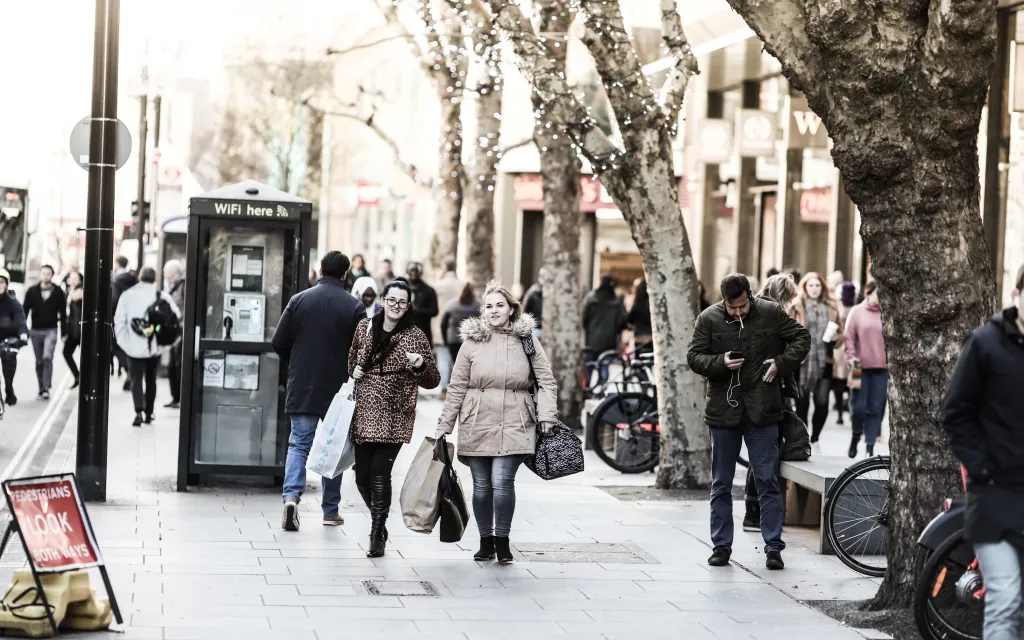A diverse group of people walking along a bustling city street lined with shops and trees.